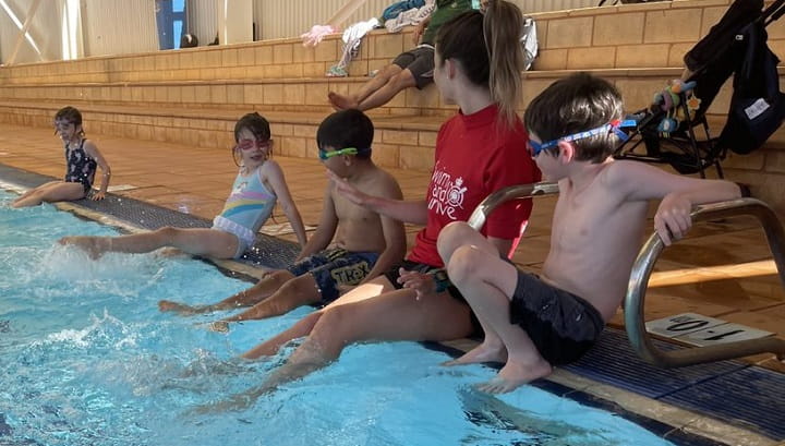 children sitting on edge of pool with their swimming teacher