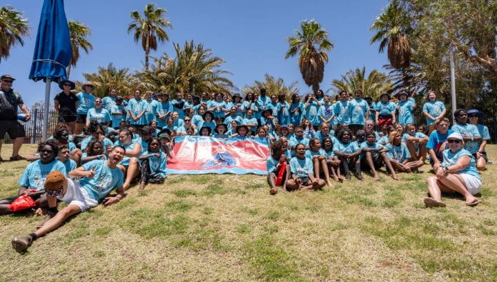 Group photo of 100 plus Aboriginal children who participated in the spirit carnival. Wearing light blue shirts with design of seagrass and turtles swimming. 