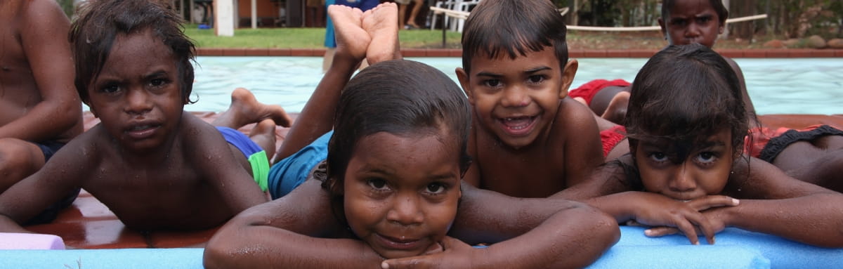 Image of 4 aboriginal boys laying on their stomachs by the side of a swimming pool smiling at camera
