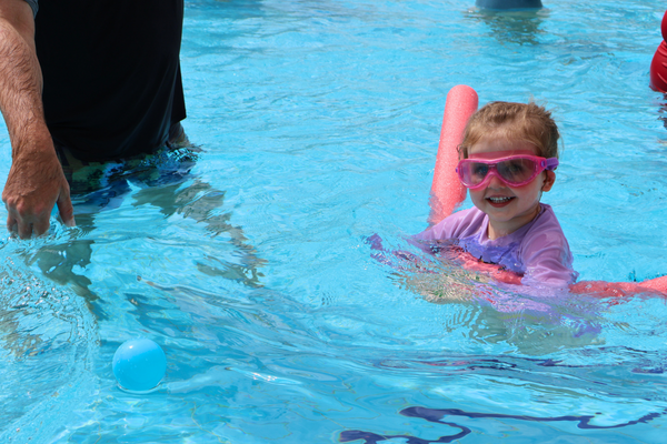 A young girl on a pool noodle smiling at the camera