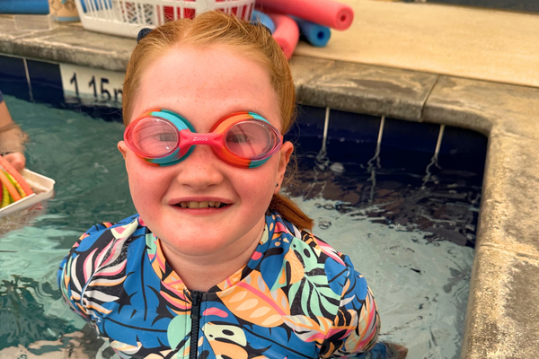 Amity, a young Swim and Survive Fund recipient, smiles up at the camera wearing goggles at the side of the pool