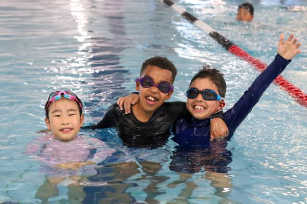 Three children smile in the pool with their arms around each other, wearing goggles during a swim session.