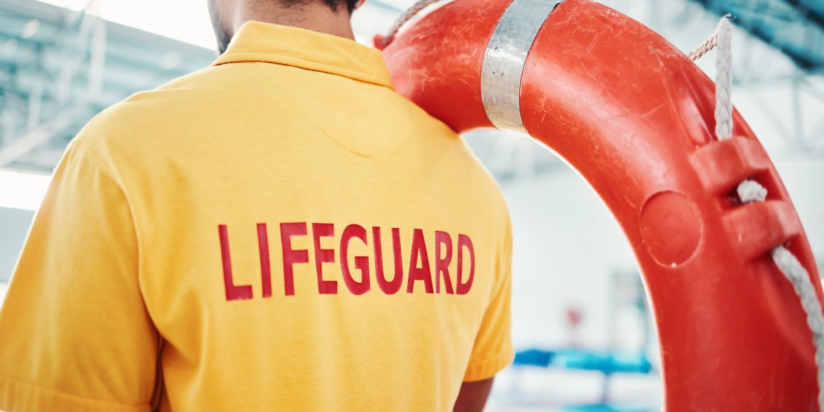 Male lifeguard wearing yellow shirt reading "lifeguard" with lifebuoy held over shoulder
