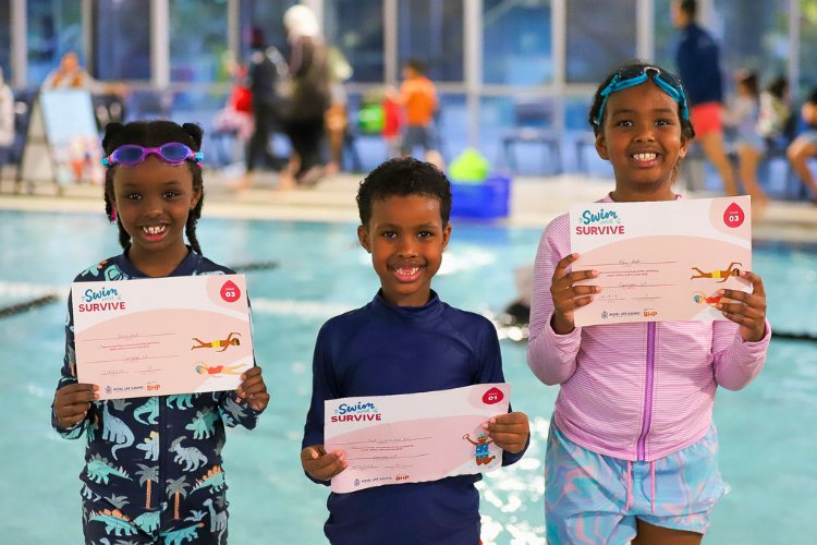 three children holding swimming stage certificates next to a pool