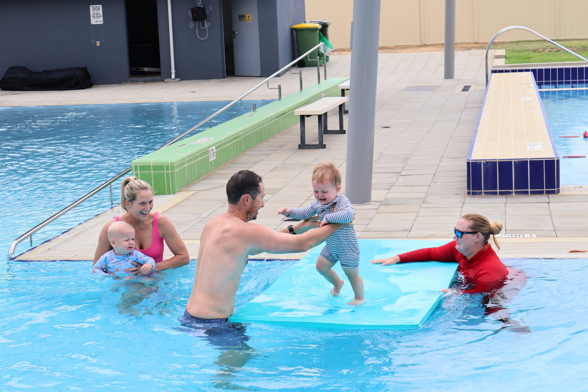 Parents hold their children in the water around a mat the instructor is holding