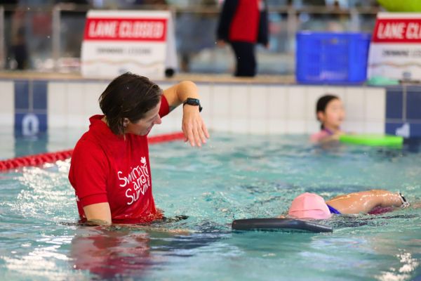 A swim instructor demonstrates an arm movement while a child practices freestyle with a kickboard in the pool.