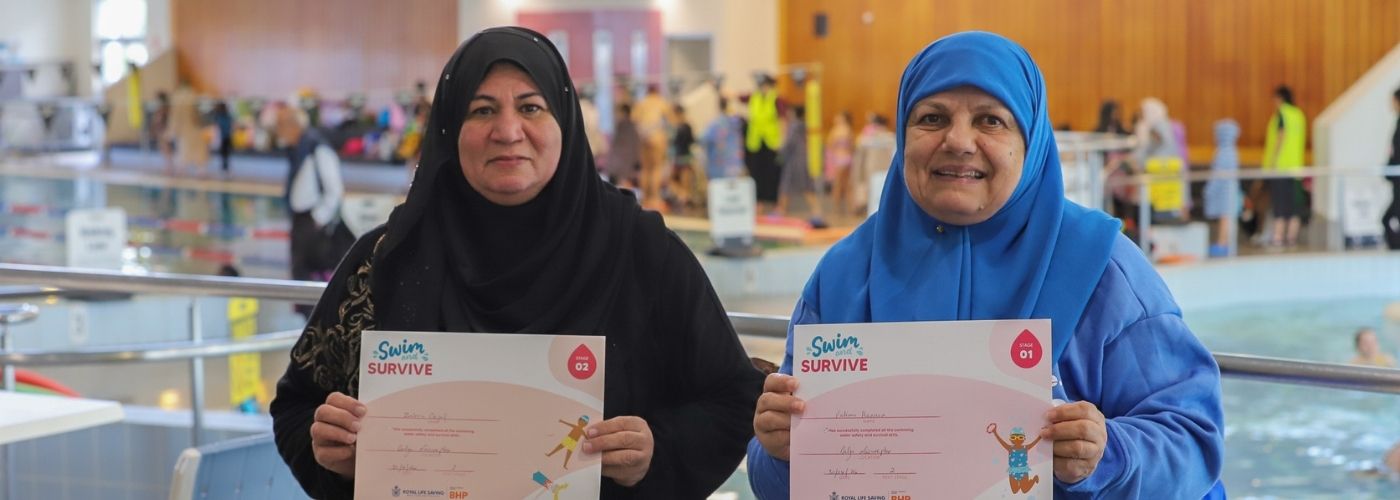 Two women wearing black and blue hijabs hold their Swim and Survive certificates beside an indoor pool.