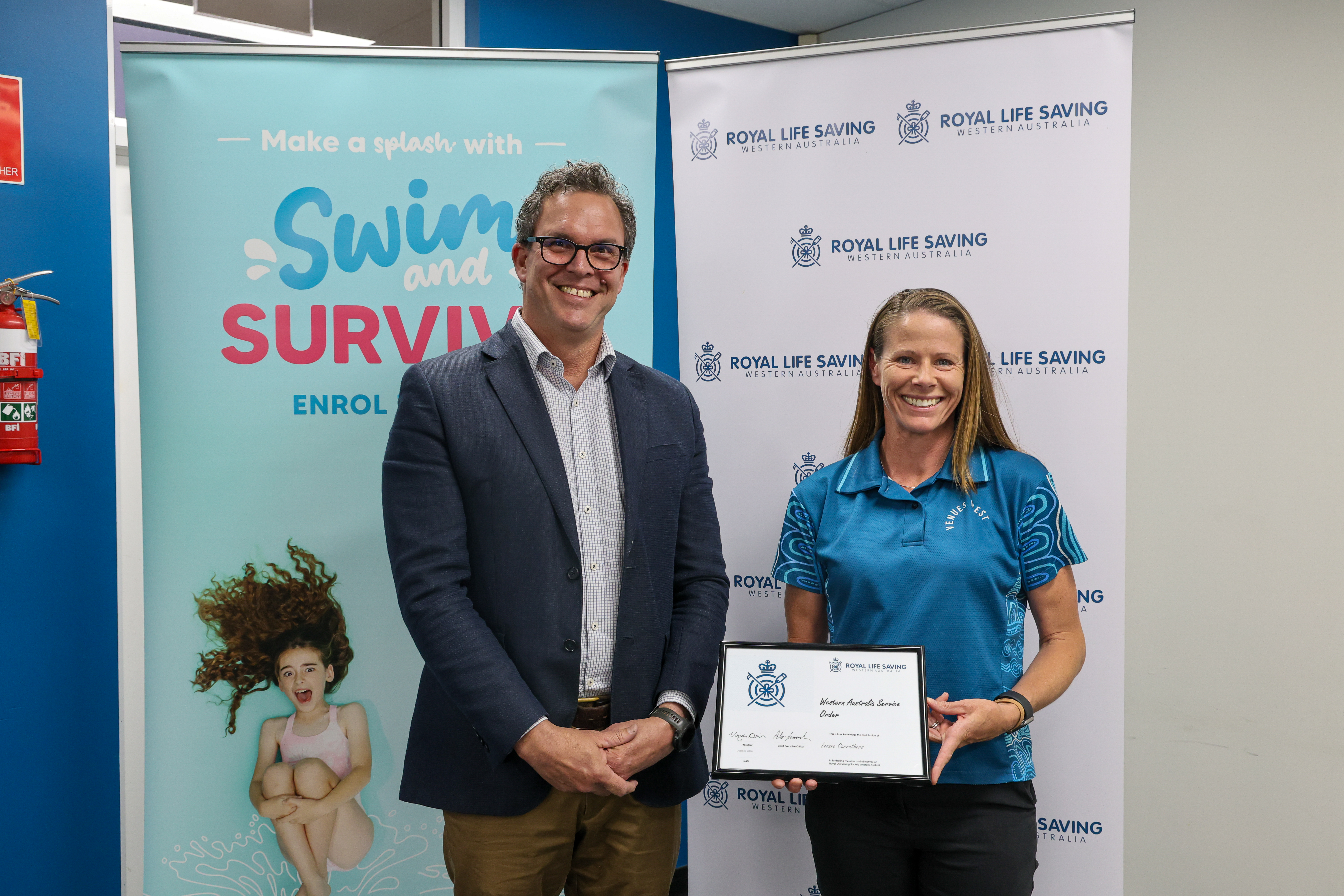 A woman holds an award certificate beside a man, both smiling in front of Royal Life Saving WA banners at the 2026 AGM.
