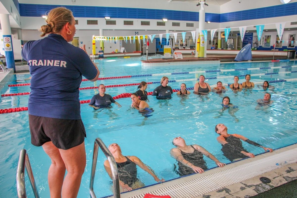 Trainer with long sleeve blue rashie standing on edge of pool instructing students in the pool