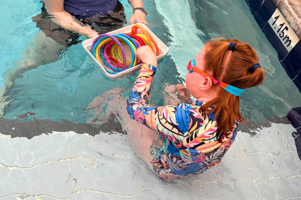 Young girl sits at the edge of the pool, looking at a bucket of toys her out of frame teacher is holding out 