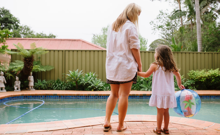 Mother and daughter holding hands looking at home pool with back towards you