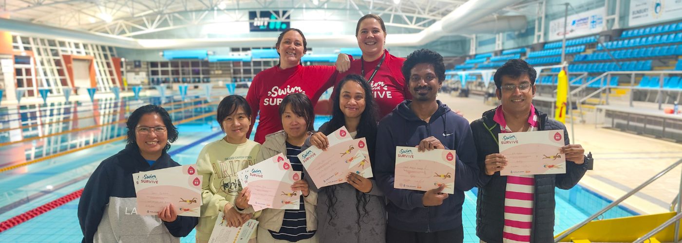 Eight smiling adults stand poolside at an indoor aquatic centre, with six participants holding swimming certificates and two instructors standing behind them in red shirts.