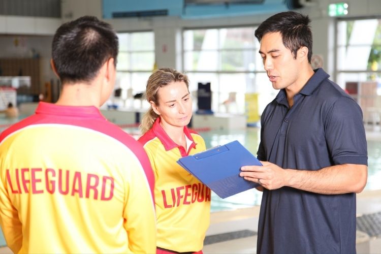 Two lifeguard trainees speaking with a supervisor holding a clipboard beside an indoor swimming pool.