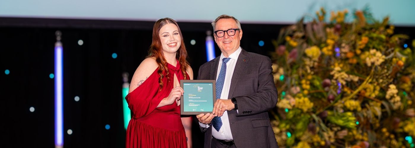  Annika Brown, wearing a red dress, holds her WA Trainee of the Year award alongside a presenter at the awards ceremony.