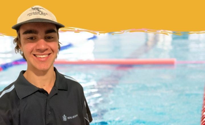 A young man wearing a khaki cap and black Royal Life Saving WA polo shirt smiles at the camera in front of an indoor swimming pool. The upper part of the image features a yellow background with a paintbrush-style border.