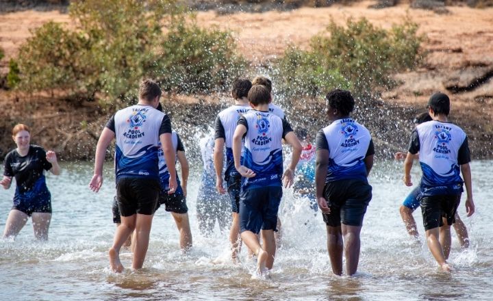 A group of young people wearing matching Talent Pool shirts walk together through a shallow body of water, splashing as they move. Bushland and red dirt are visible in the background.