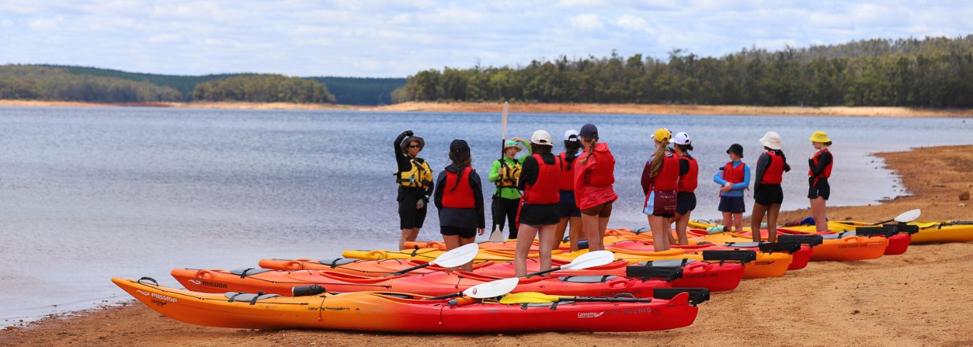 Students stand wearing lifejackets with backs to camera while the trainer speaks with kayaks at their feet