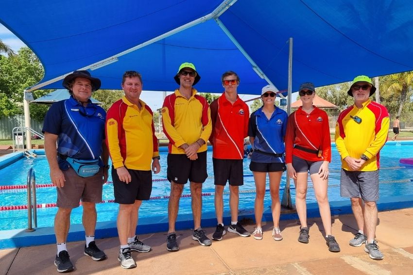 Group of 7 lifeguards and pool managers standing in a row smiling towards camera with pool in background