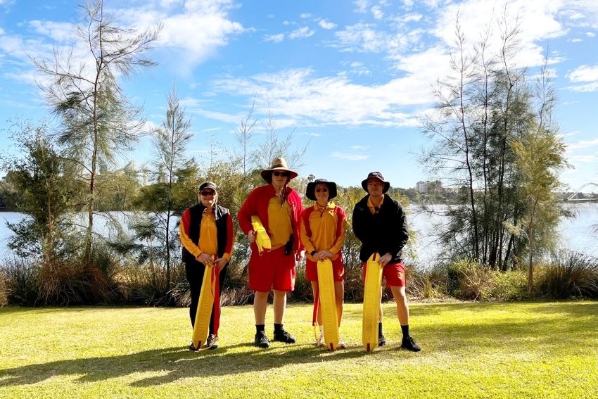 Four lifeguards, dressed warmly, standing in a row posing for photo with the swan river in the background