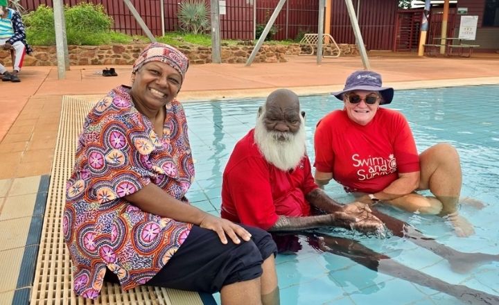 Three smiling adults sit at the edge of a swimming pool, with one man partially in the water holding hands with a swim instructor in a red