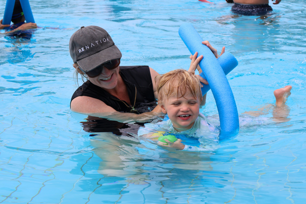 A young boy on a pool noodle smiles at the camera with his mum behind him