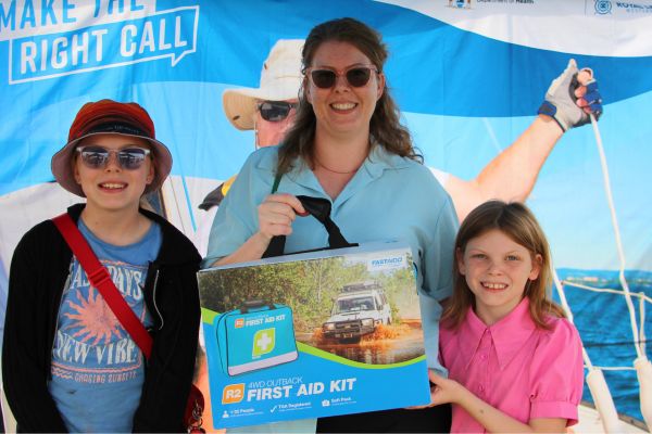 A smiling woman and two children hold an emergency first aid kit prize in front of a Make the Right Call banner at the FeNaClNG Festival.