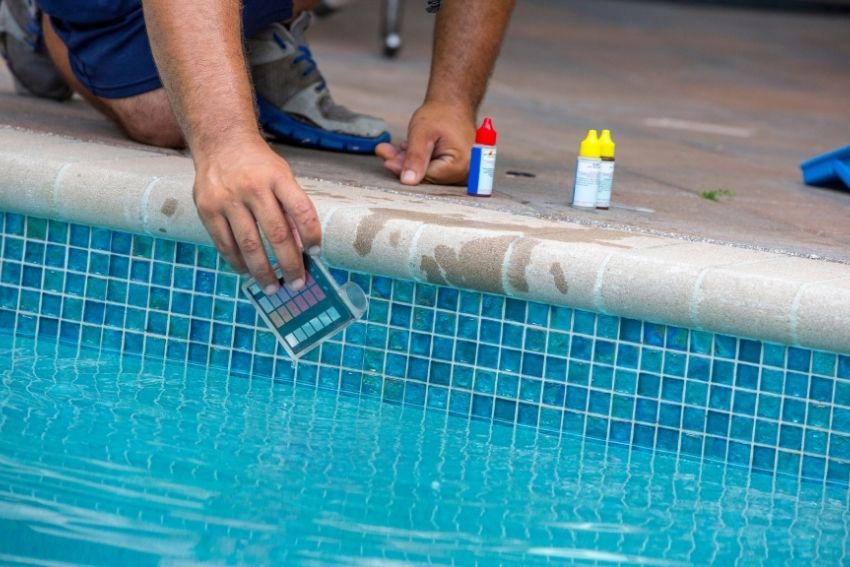 Close of up the edge of a home pool with person kneeling next to it scooping water out with testing materials to test