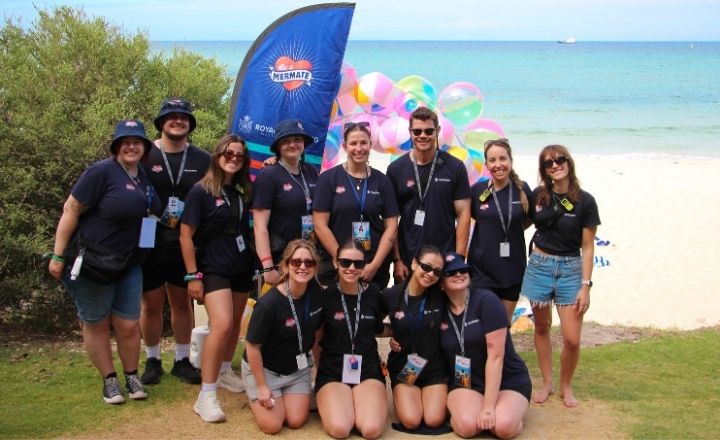 A group of Royal Life Saving WA volunteers wearing matching navy event shirts and lanyards pose in front of a colourful beach ball display and ocean backdrop during a beach event.