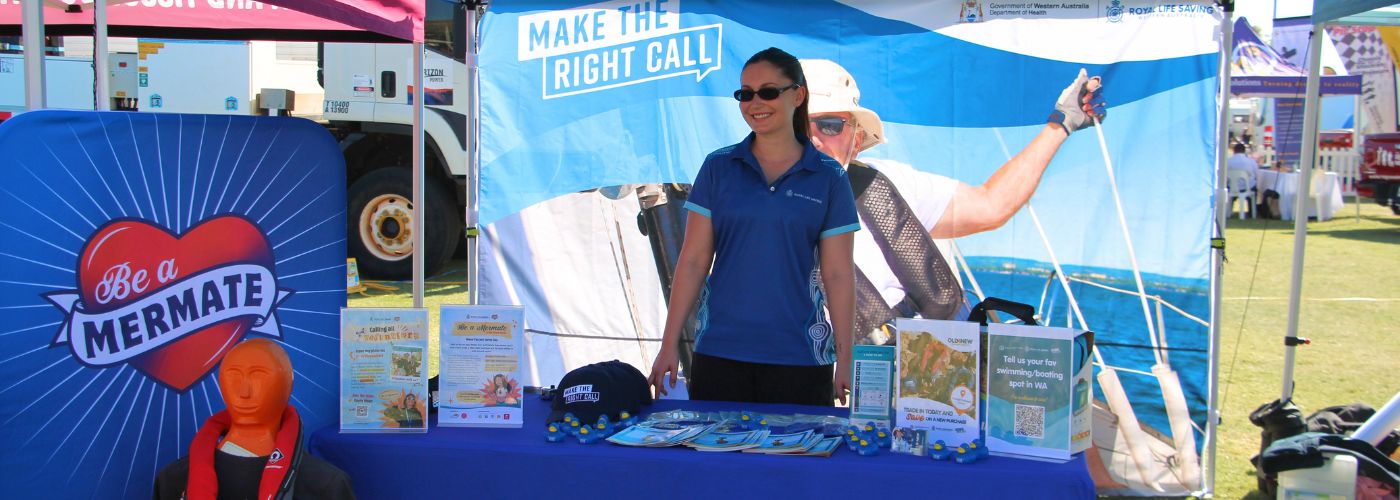 A Royal Life Saving WA staff member stands behind a water safety information stall featuring Be a Mermate and Make the Right Call displays at the FeNaClNG Festival.