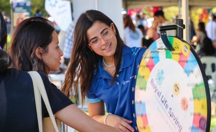 A young woman in a Royal Life Saving polo shirt helps another woman spin a colourful prize wheel at a community event booth, with people and tents visible in the background.