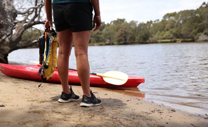 A person standing on a sandy riverbank holding a yellow life jacket, facing a red kayak and paddle resting at the water’s edge. Trees and calm water are visible in the background.