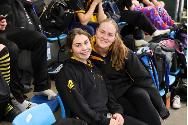 Two Western Australian athletes smile together while seated in the spectator stands.