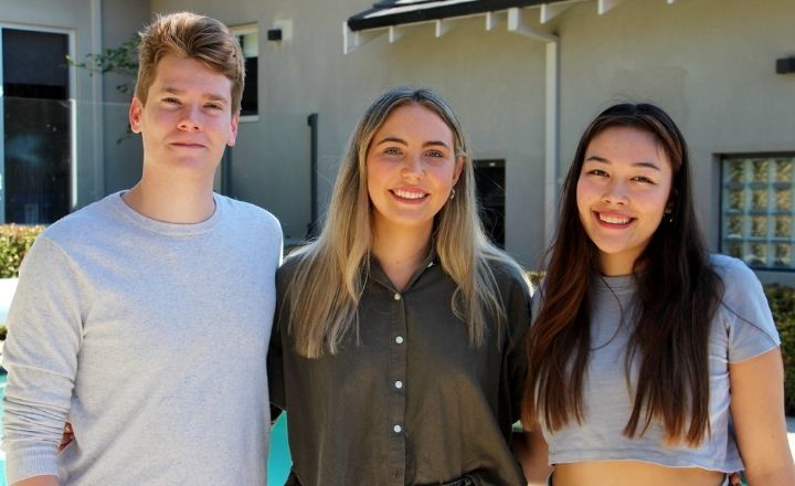 Three young adults stand side by side outdoors, smiling at the camera. Behind them is a pool and the exterior of a modern building.