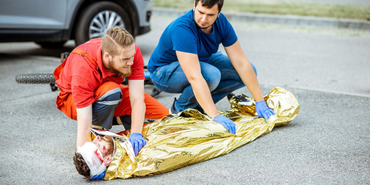 Person lying in the road bleeding with a metallic blanket and two bystanders holding on to help