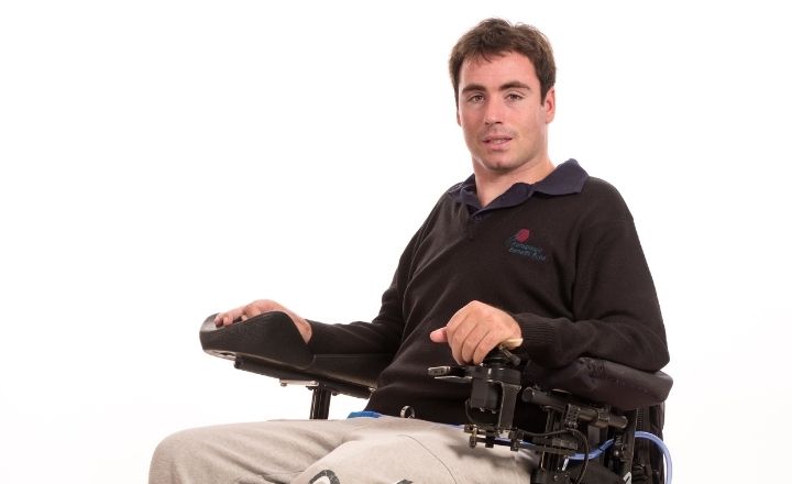 Portrait of Mathew Naysmith seated in a powered wheelchair against a white background, wearing a black polo shirt and grey pants, looking directly at the camer