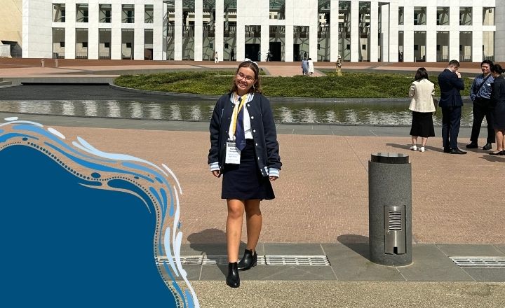 Young woman smiling and standing in front of Parliament House wearing a badge and jacket.