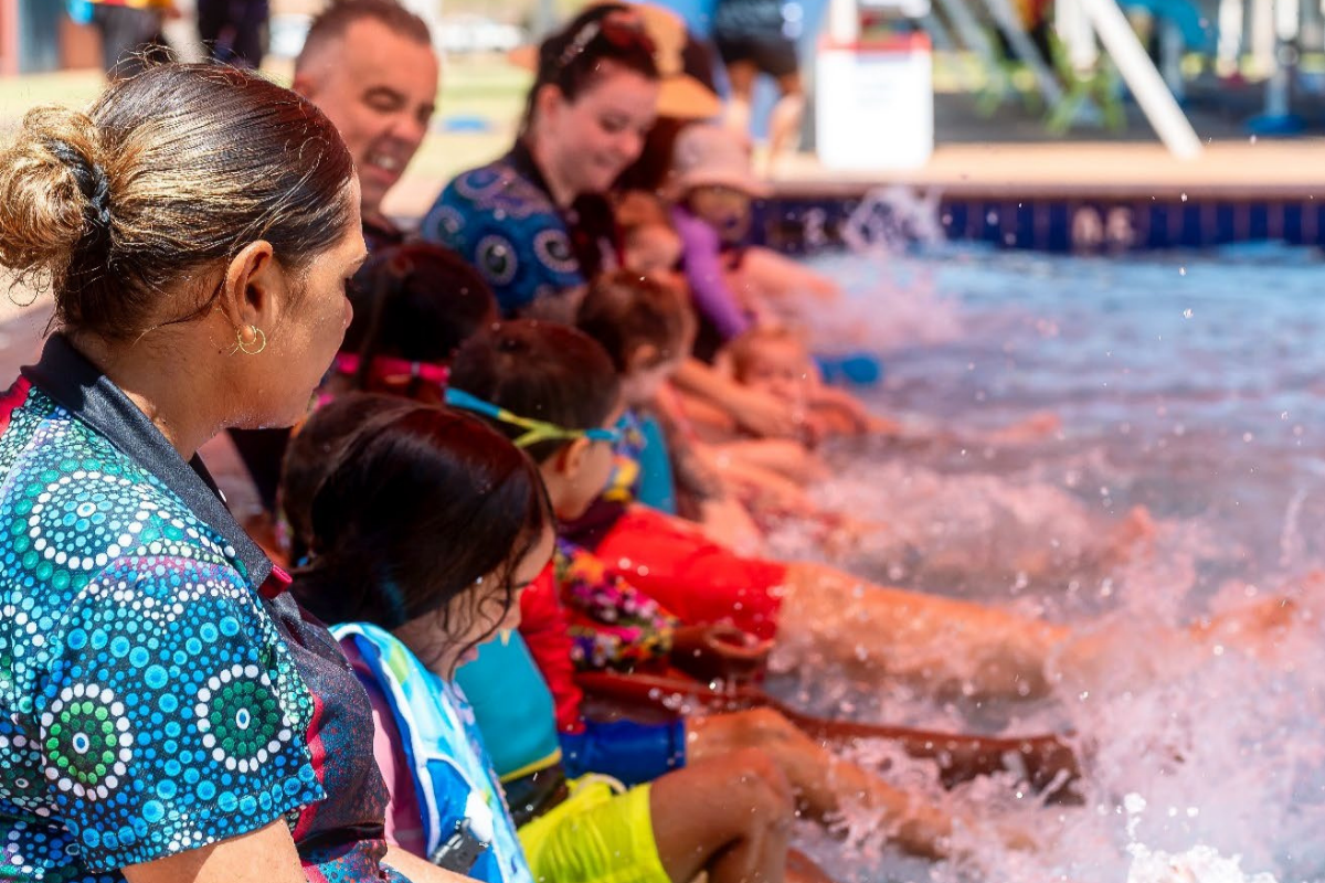 Line of parents and children sit along pool edge