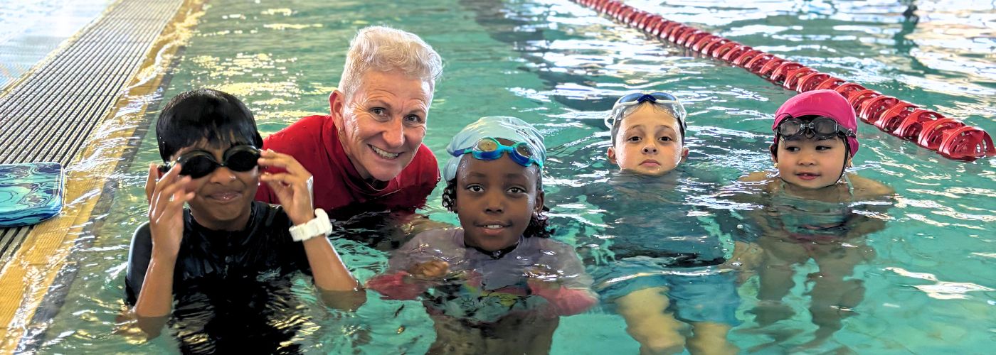 A swim instructor and students face the camera while standing submerged in the water
