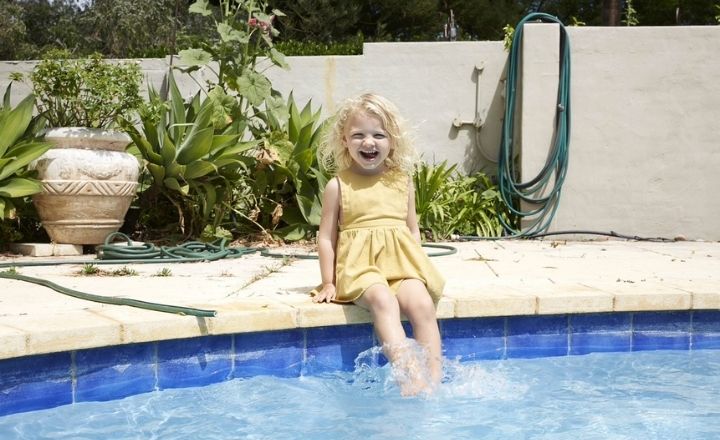 3 year old girl with blonde curly hair in yellow dress sitting on edge of pool and laughing at camera