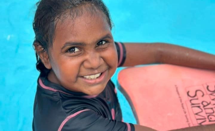 A young girl smiles brightly while holding a pink kickboard in a swimming pool. She wears a dark rash shirt with pink trim, and the kickboard features the “Swim and Survive” logo.