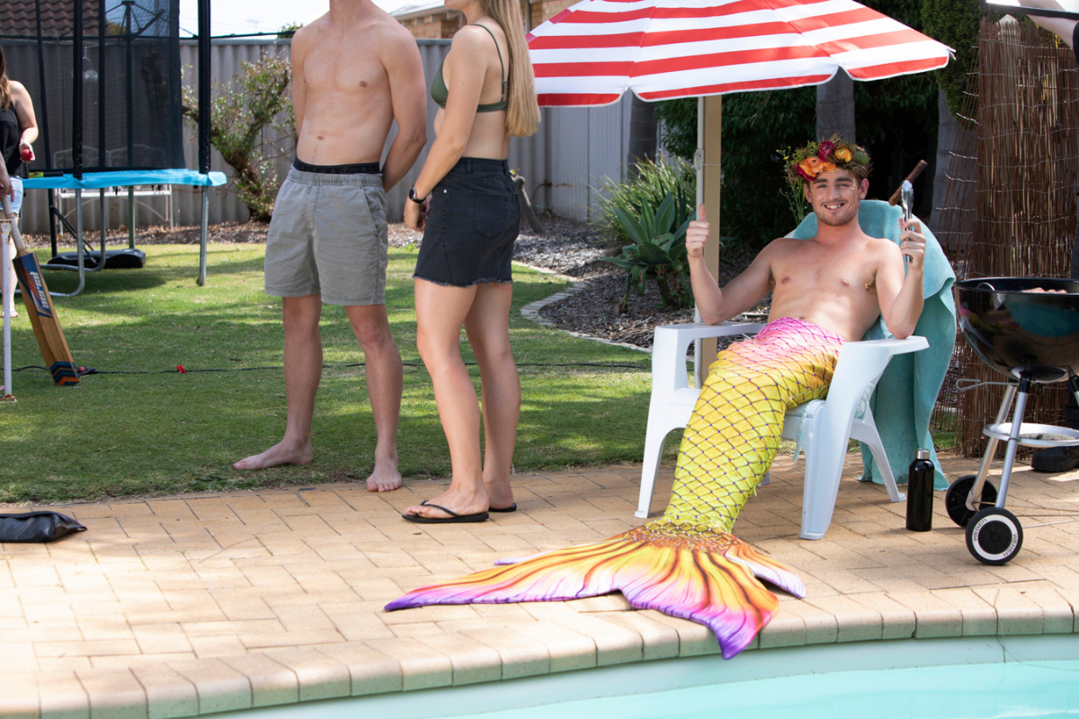 Man in mermaid tail sits under umbrella in a backyard