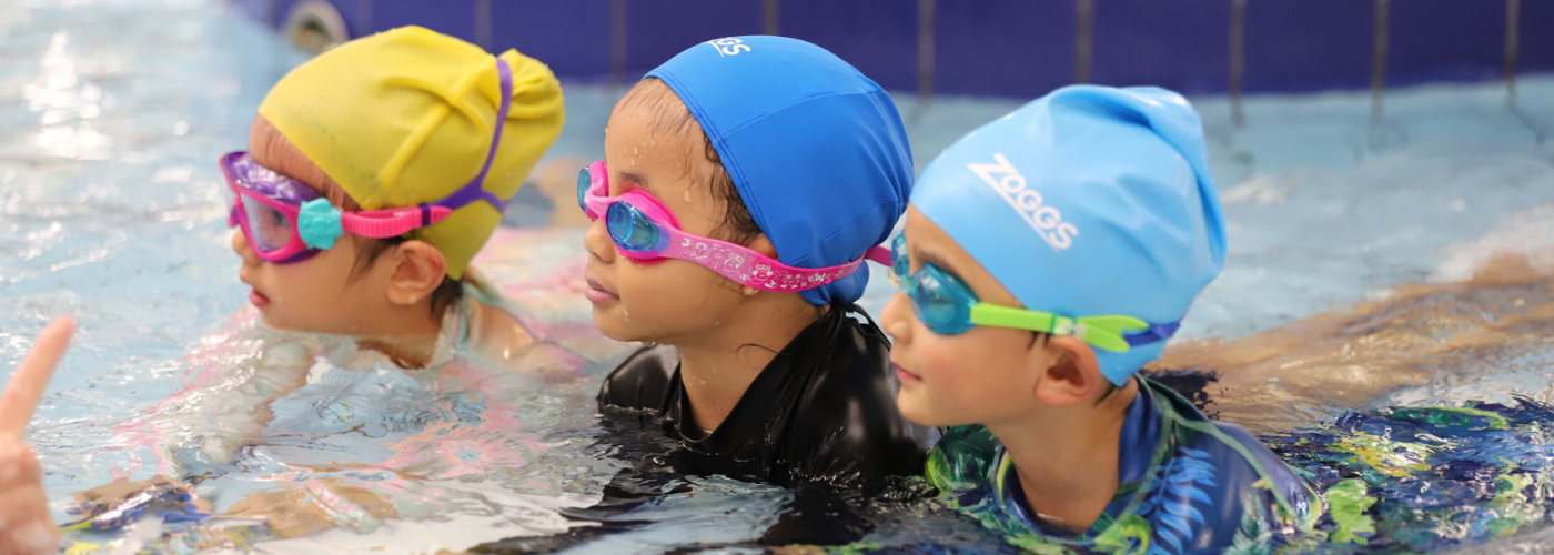 Three children in swimming caps and goggles lay on their stomachs in shallow water
