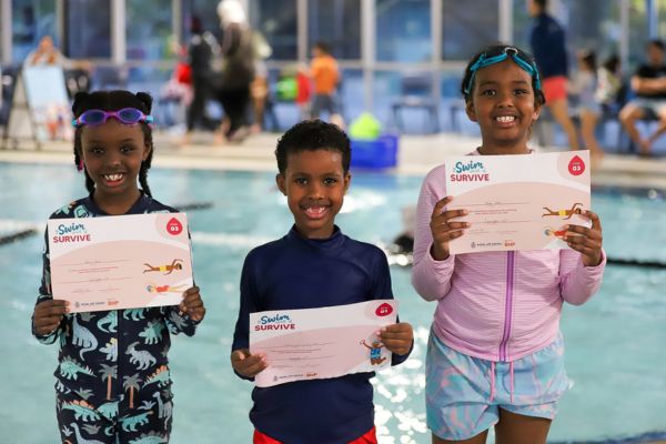 Three children stand at the pool edge holding swim program certificates and smiling at the camera.