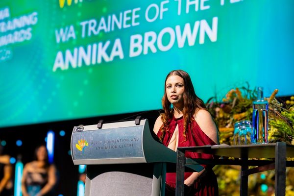 Annika Brown speaks at a lectern, with a screen behind her announcing her as WA Trainee of the Year.