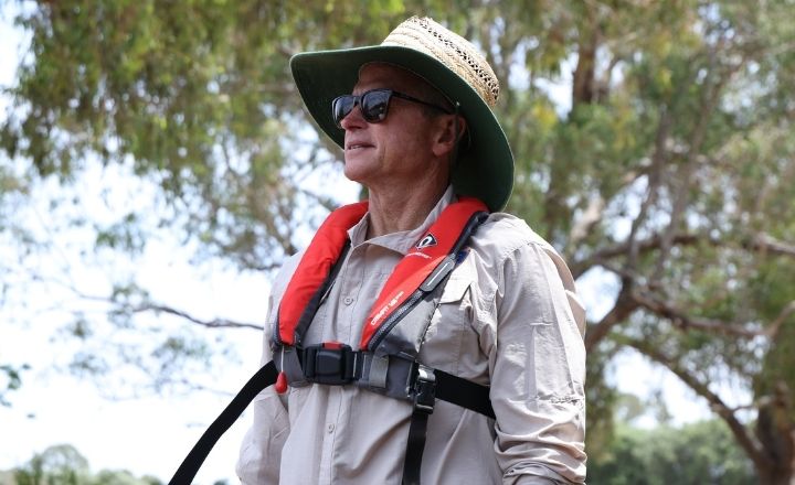 Close up of caucasion man in his fifties wearing a slim profile lifejacket over his beige fishing shirt. Also wearing sunglasses and a wide brim hat
