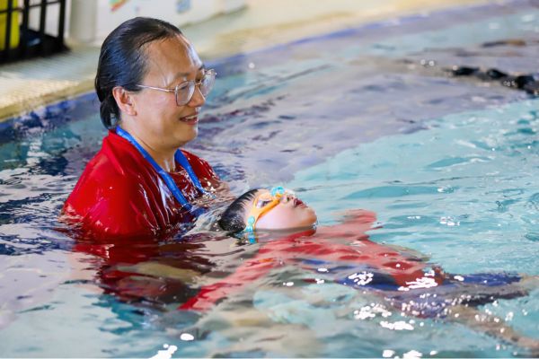 An adult swim instructor supports a child floating on their back in an indoor swimming pool during a lesson.