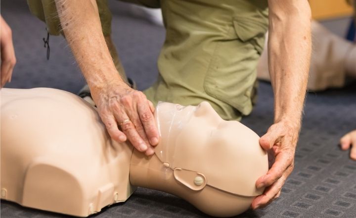 Close up of profile of cpr manikin laying on the floor with a older persons knee next to it wearing light green hiking pants.