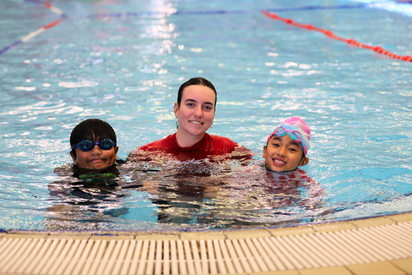 A swim teacher stands between two students in shallow water, all smiling at the camera