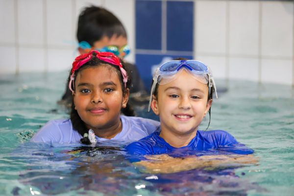 Two children wearing goggles smile while standing close together in the pool during a swimming lesson.