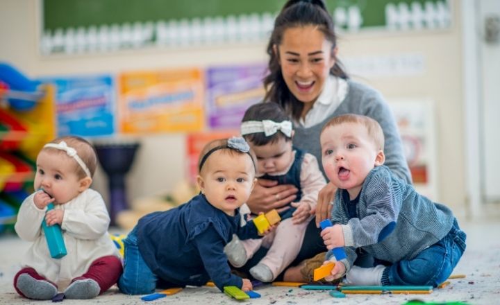 daycare worker fuzzy in the background sitting on the floor playing with crawling infants who are in focus. All are smiling and laughing